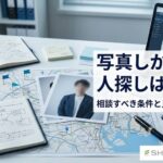 机の上の人物写真や地図、メモ、ノートPCを使って、少ない手がかりから人探しの情報を整理しているイメージ